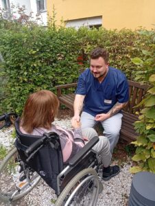 Robin Krüger sitzt im Garten des AWO Seniorenzentrums Lotte Lemke in Bad Kreuznach auf der Bank. Er hält die Hand einer im Rollstuhl sitzenden Bewohnerin, die man nur von Hinten sieht. Hinter und neben der Bank wächst eine grüne Hecke.
