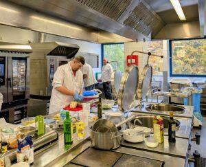 Zu sehen ist die Küche des Seniorenzentrums Lotte Lemke in Bad Kreuznach. Im Vordergrund ein Herd mit Töpfen und Zutaten wie Öl und Salz für die Zubereitung der Gerichte. Im Hintergrund bereiten drei Teilnehmer*innen des Kochworkshops die Speisen zu.