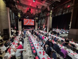 Luftaufnahme des Konferenzraums während der Bundeskonferenz der AWO. Der Raum ist mit mehreren langen Tischen ausgestattet, die in einer geraden Reihe angeordnet sind. Die Tische sind ordentlich gedeckt, und es ist eine große Anzahl an Stühlen darum herum angeordnet. Die Atmosphäre wirkt professionell und organisiert, während die Teilnehmer vermutlich in angeregten Gesprächen oder Diskussionen vertieft sind. Im Hintergrund ist eine Präsentationswand oder ein Bildschirm sichtbar, der möglicherweise für die Veranstaltung genutzt wird.