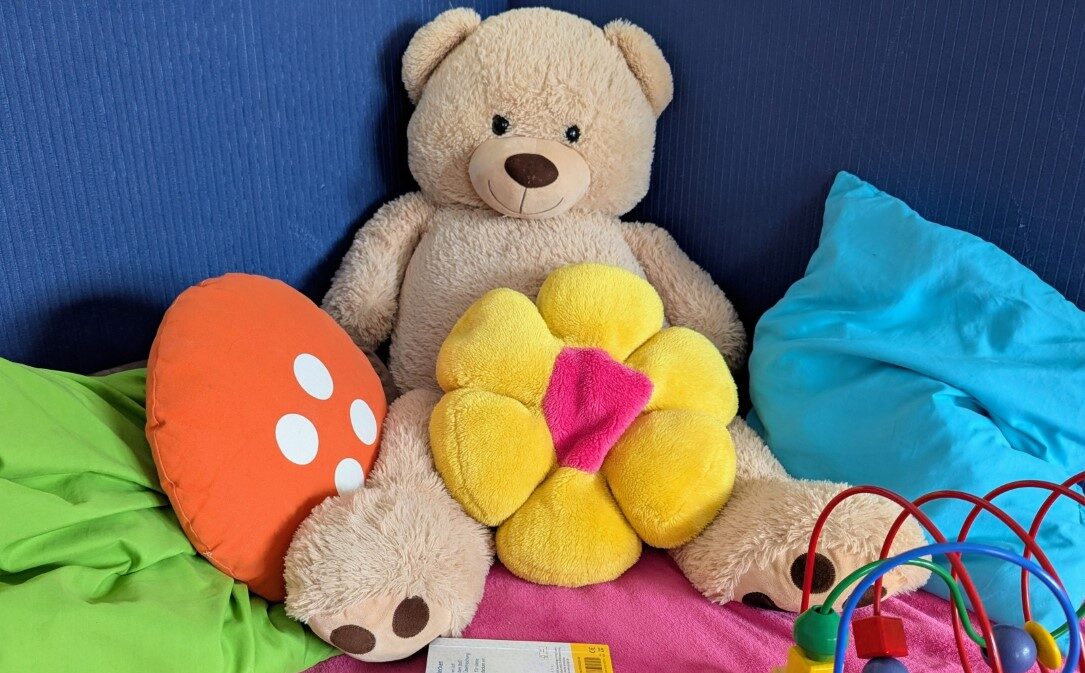 Ein Teddybär lehnt an der Wand. Links und rechts liegen bunte Kissen in grün und blau. Eins davon hat die Form einer Sonne. Kindertagespflegestelle