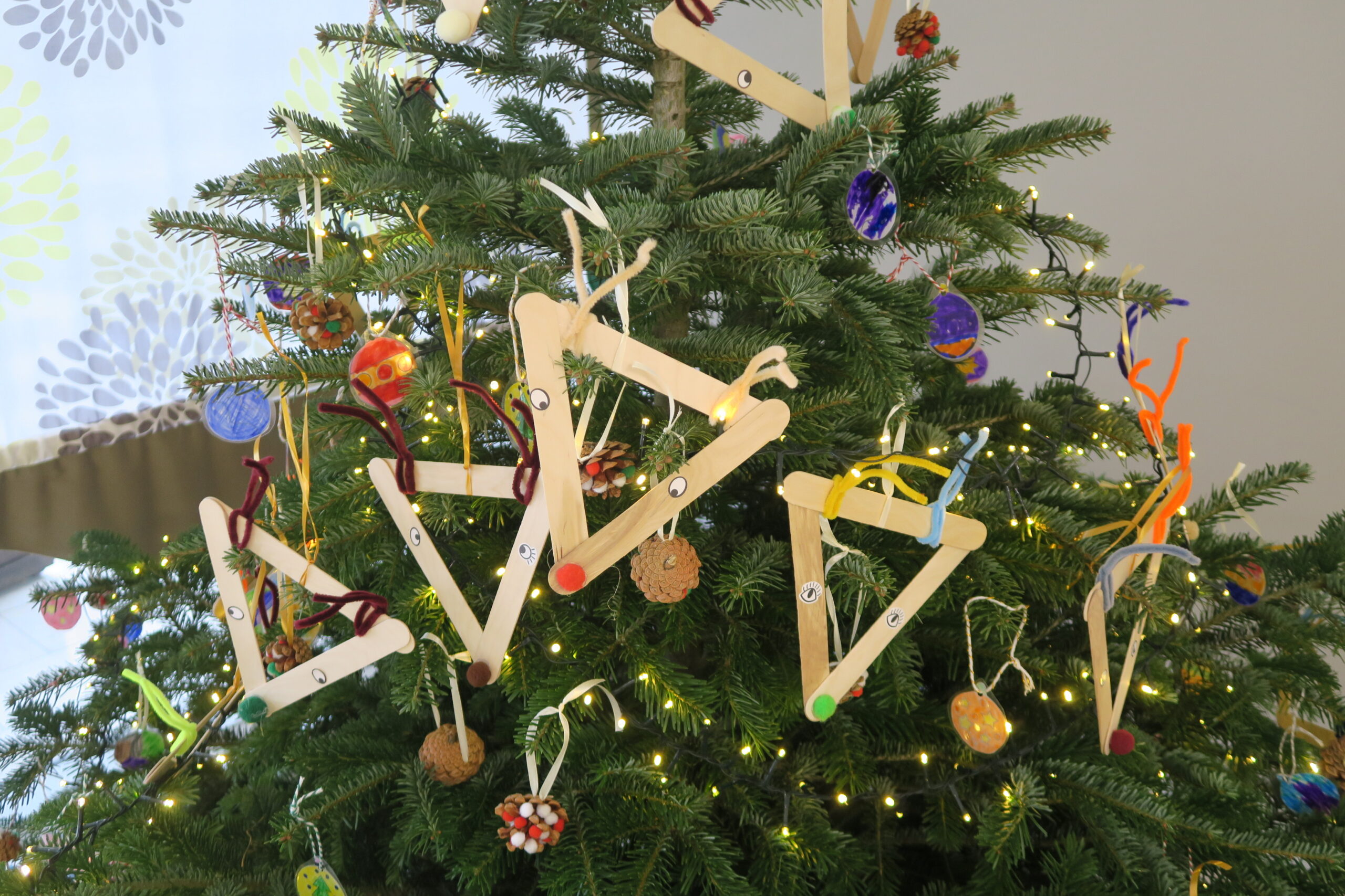 Das Bild zeigt eine Tanne. Sie ist geschmückt mit Holzdreiecken, Tannenzapfen und bunt gebastelten Papierschmuck. Ringsherum ist eine Lichterkette. Der Baum steht in der Eingangshalle des Seniorenzentrums. Baumschmücken für Groß und Klein