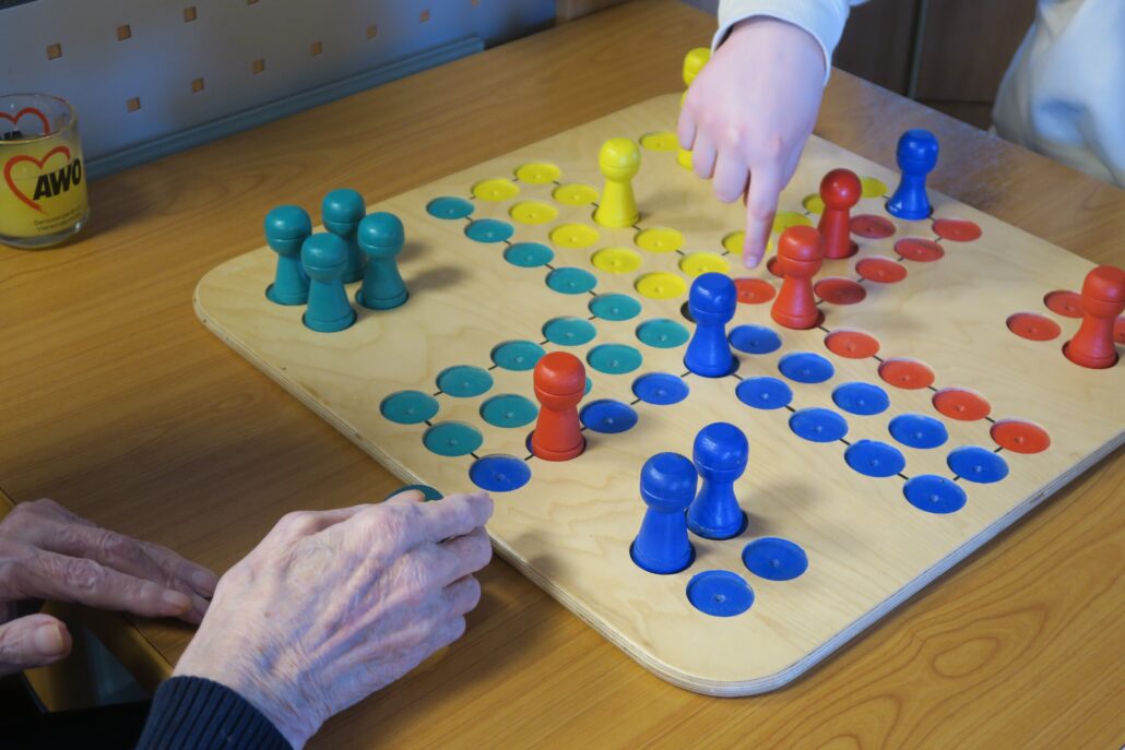 Das Bild zeigt ein buntes Spiel aus Holz, Mensch ärgere dich nicht. Die Figuren haben die Farben blau, grün, gelb und rot. 6 Spielfiguren sind bereits im Spiel. Zu sehen ist eine Kinderhand und die Hände eines Senior.