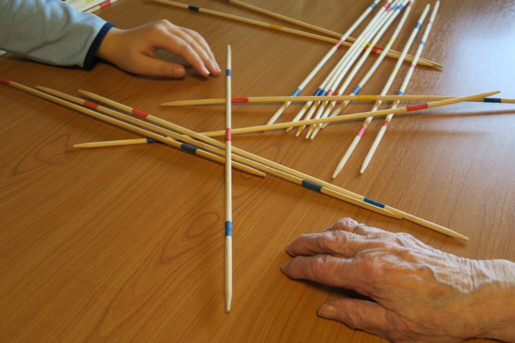 Spielevormittag: Das Bild zeigt die Stäbe eines Mikadospiel, die kreuz und quer übereinander liegen. Zu sehen ist eine Kinderhand und die Hand einer Seniorin.
