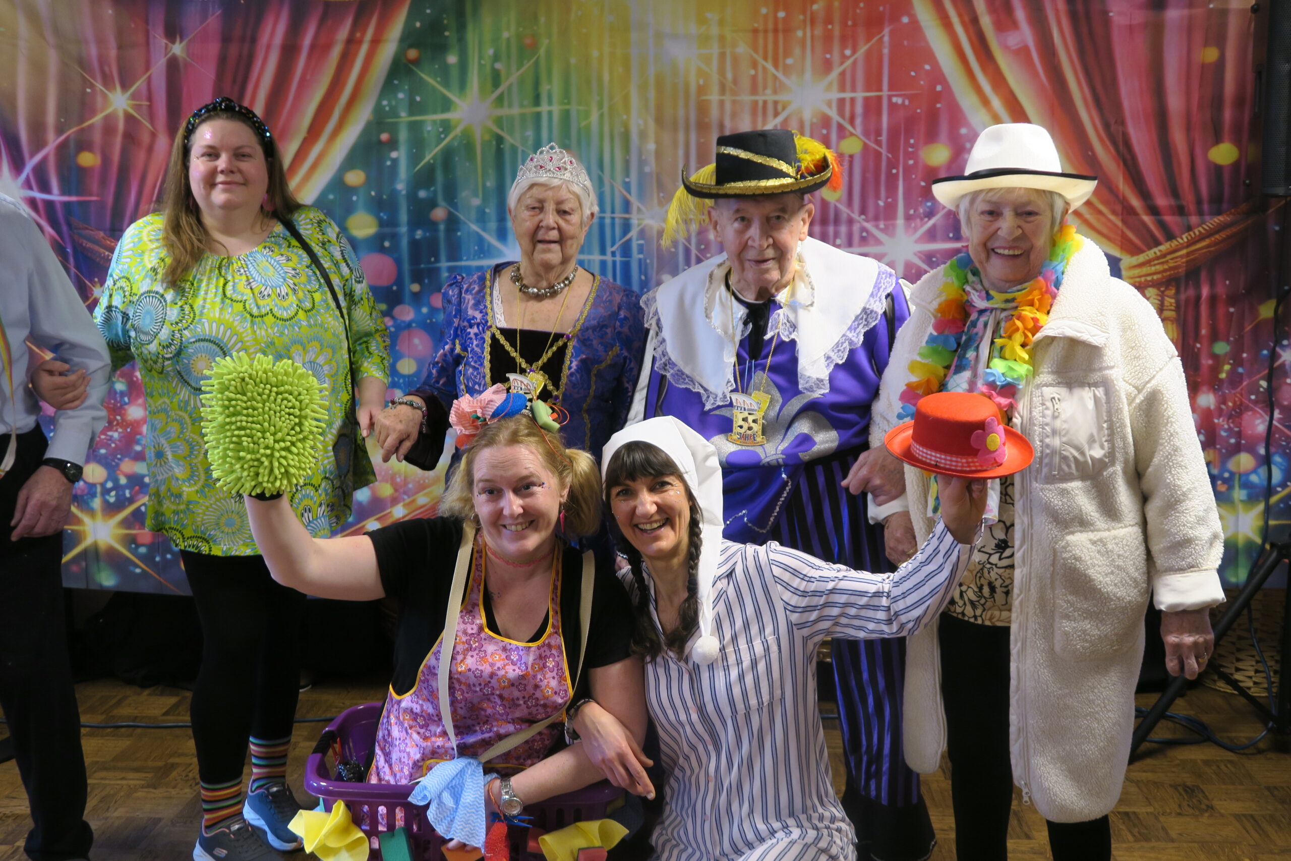 Vier Personen unterschiedlicher Karnevalsgesellschaften stehen in verschiedenen Kostümen verkleidet vor einem bunten Hintergrund. Davor hocken zwei ebenfalls verkleidete Frauen und Halten die Hände hoch. Alle abgebildeten Personen lachen.