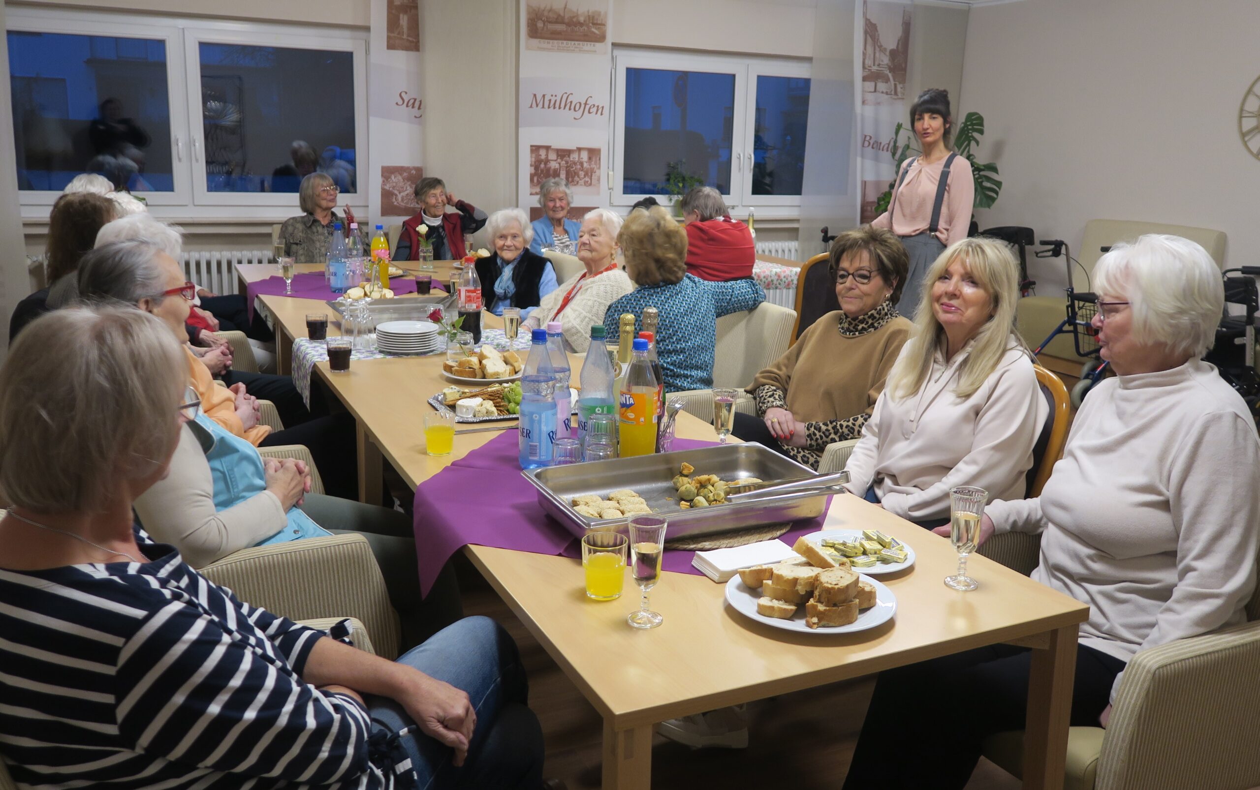Das Bild zeigt einen gedeckten Tisch, Käseplatte, Blätterteigtaschen, Softgetränke, Sektflaschen für den Frauenstammtisch. Drumherum sitzen Frauen. Sie lachen und sprechen miteinander. Einige Lächeln in die Kamera.