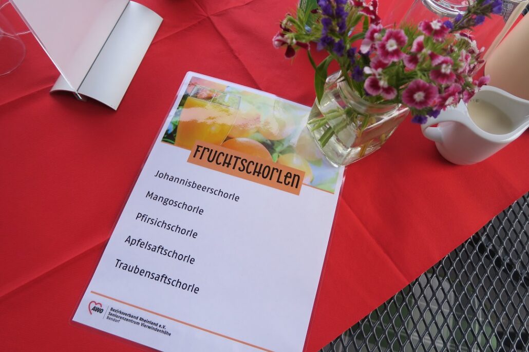 Eine Getränkekarte auf der verschiedene Fruchtschorlen abgedruckt sind, liegt auf einem Tisch. Daneben steht ein Blumenstrauß.