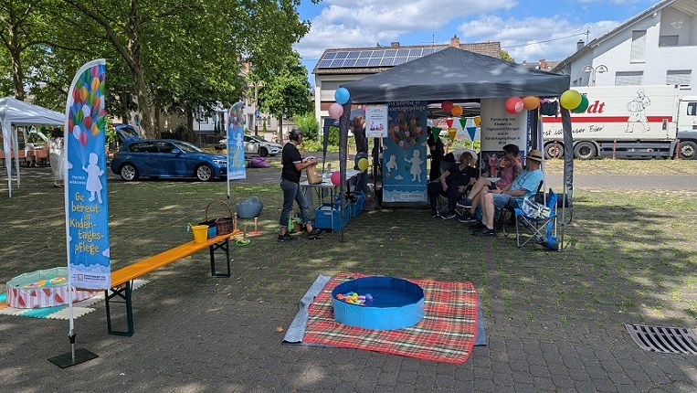 Ein Pavillon, unter dem Menschen sitzen. Links stehen Tische mit Informationsmaterial. Vorne im Bild steht ein Planschbecken.