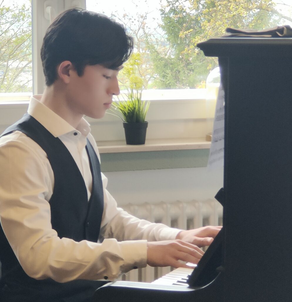 Das Bild zeigt einen jungen Pianist am klavier. Er trägt ein weißes Hemd und eine schwarze Weste. Das Klavier ist schwarz und stammt aus vergangenen Zeiten. Der Mann bespielt das Klavier.
