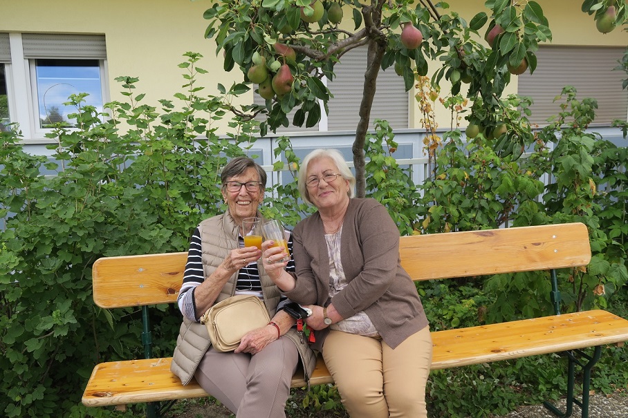 Zwei Frauen sitzen auf einer Bank unter einem Birnbaum mit einem Getränk in der Hand. Sie lachen in die Kamera.