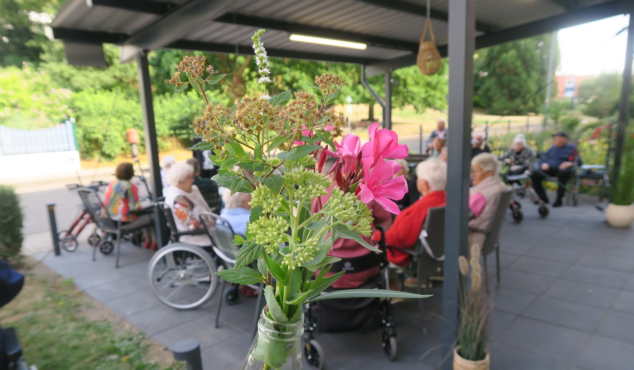 Im Vordergrund ist ein grün-pinker Blumenstrauß zu sehen. Im Hintergrund sitzen Menschen unter einem Pavillon in der Vierwindenhöhe.