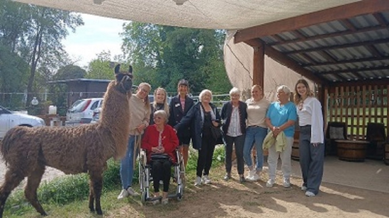 Auf dem Bild links ist ein braunes Lama zu sehen. Neben ihm stehen 8 Frauen und eine Frau sitzt in einem Rollstuhl. In allen Gesichtern ist ein Strahlen zu sehen.