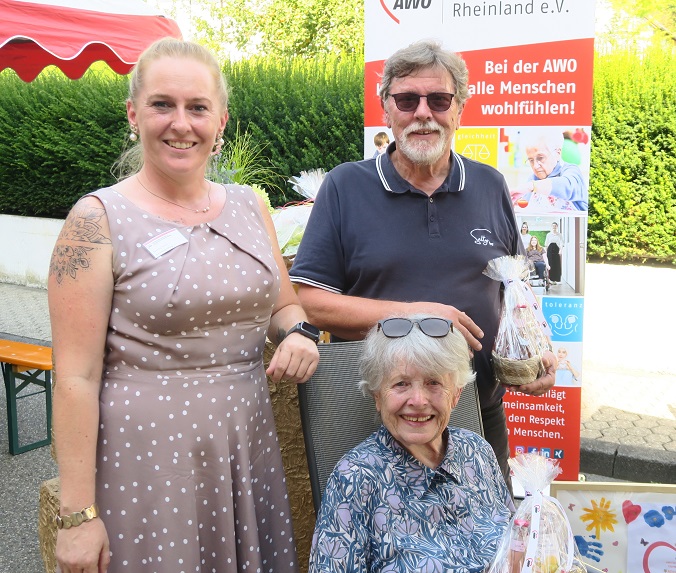 Eine ältere Frau sitzt auf einem Stuhl und lächelt in die Kamera. Dahinter stehen links eine Frau und rechts ein Mann, die ebenfalls in die Kamera lächeln.