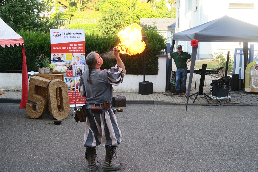 Ein Feuerspucker spuckt eine Flamme. Im Hintergrund steht ein Mann unter einem Pavillon und eine goldene 50 dekoriert ein Rednerpult.