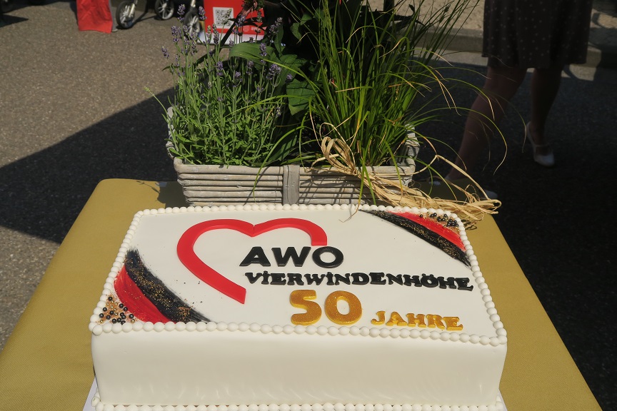 Auf einem Tisch steht im Vordergrund eine Torte mit weißer Creme. Auf der Torte ist das Herz-Logo der AWO mit dem Zusatz "AWO Vierwindenhöhe 50 Jahre" abgebildet.