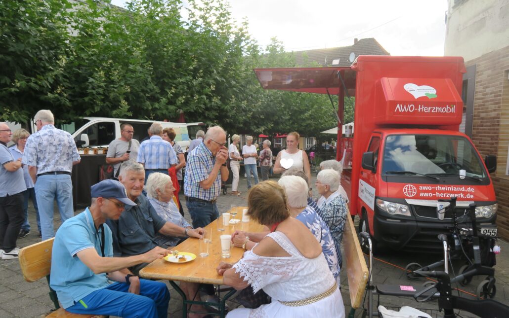 Das Bild zeigt viele Menschen. Einige sitzen im Vordergrund zusammen an einer Biertischgarnitur und schauen zu einem Mann, der am Tisch steht und Mundharmonika spielt. Im Hintergrund ist das rote AWO Herzmobil zu sehen. Menschen stehen zusammen und unterhalten sich.