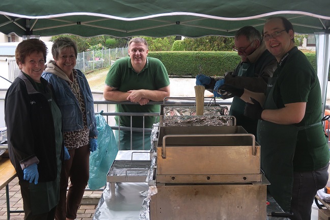 Unter einem Pavillon stehen vier Menschen und blicken in die Kamera. In der Mitte stehen große Pfannen, auf denen Backfisch gebacken wird.
