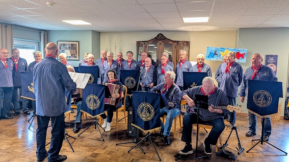 Männer in Seemannstracht stehen als Chor formiert und singen. Vorne sitzen fünf Personen, die verschiedene Instrumente spielen.