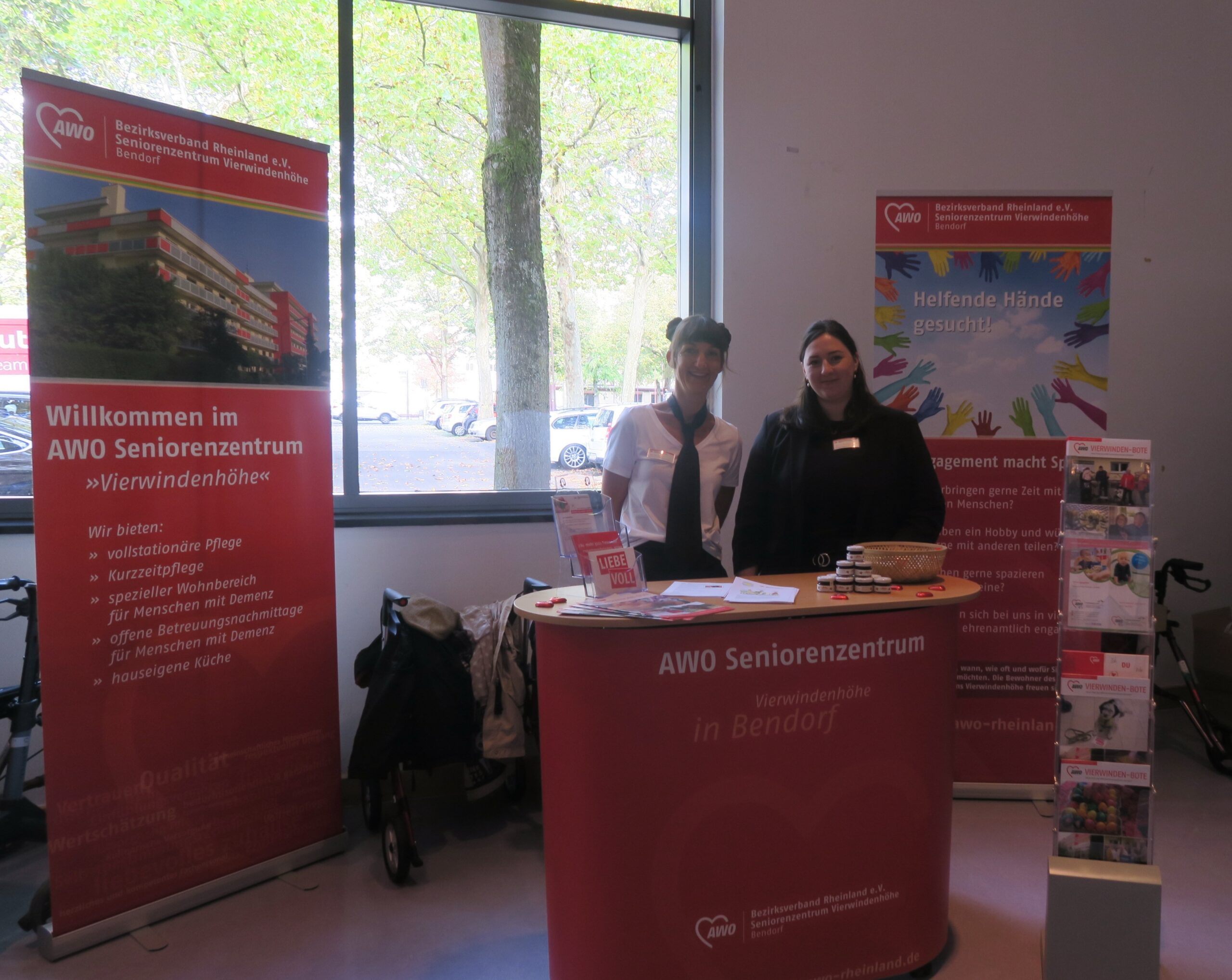 Das Bild zeigt den Stand des AWO Seniorenzentrum. Vorne im Bild ein roter Tisch, dahinter stehen zwei Mitarbeiterinnen der Einrichtung. Hinten rechts und links im Bild zwei Rollups mit Informationen und Bildern über das Seniorenzentrum.