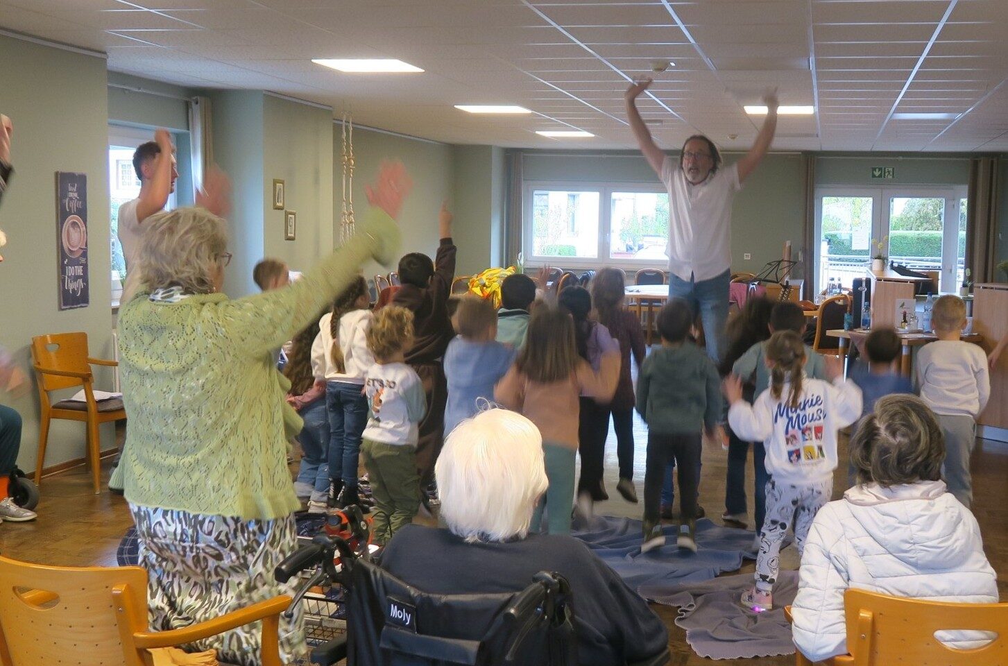 Das Bild zeigt Senior*innen und Kinder, die gemeinsam in Bewegung sind. Eine Dame streckt beide Arme weit nach oben, Kinder springen und der Märchenpädagoge Herr Meyer hebt beide Arme und springt nach oben.